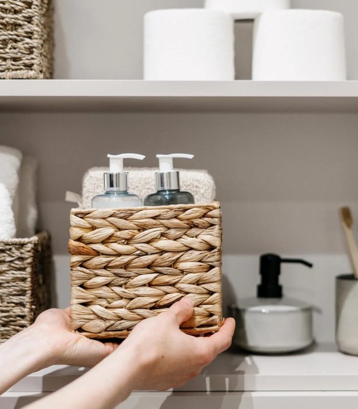 Organization of space in the bathroom cabinet. Cropped view of woman putting wicker box with bath sponge, shampoo, soap dispenser bottle and other cosmetics products in closet